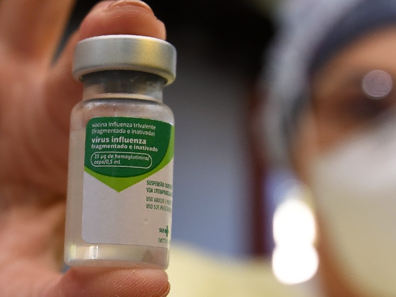 Foto do frasco da vacina contra Influenza em primeiro plano, segurado por uma profissional da saúde que aparece ao fundo com parte do rosto desfocado. O rótulo do frasco é branco e verde e traz informações técnicas sobre o imunizante